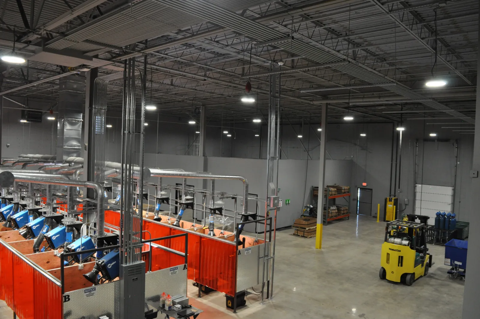 Industrial warehouse interior with welding stations, bright overhead lights, and a yellow forklift on concrete floor