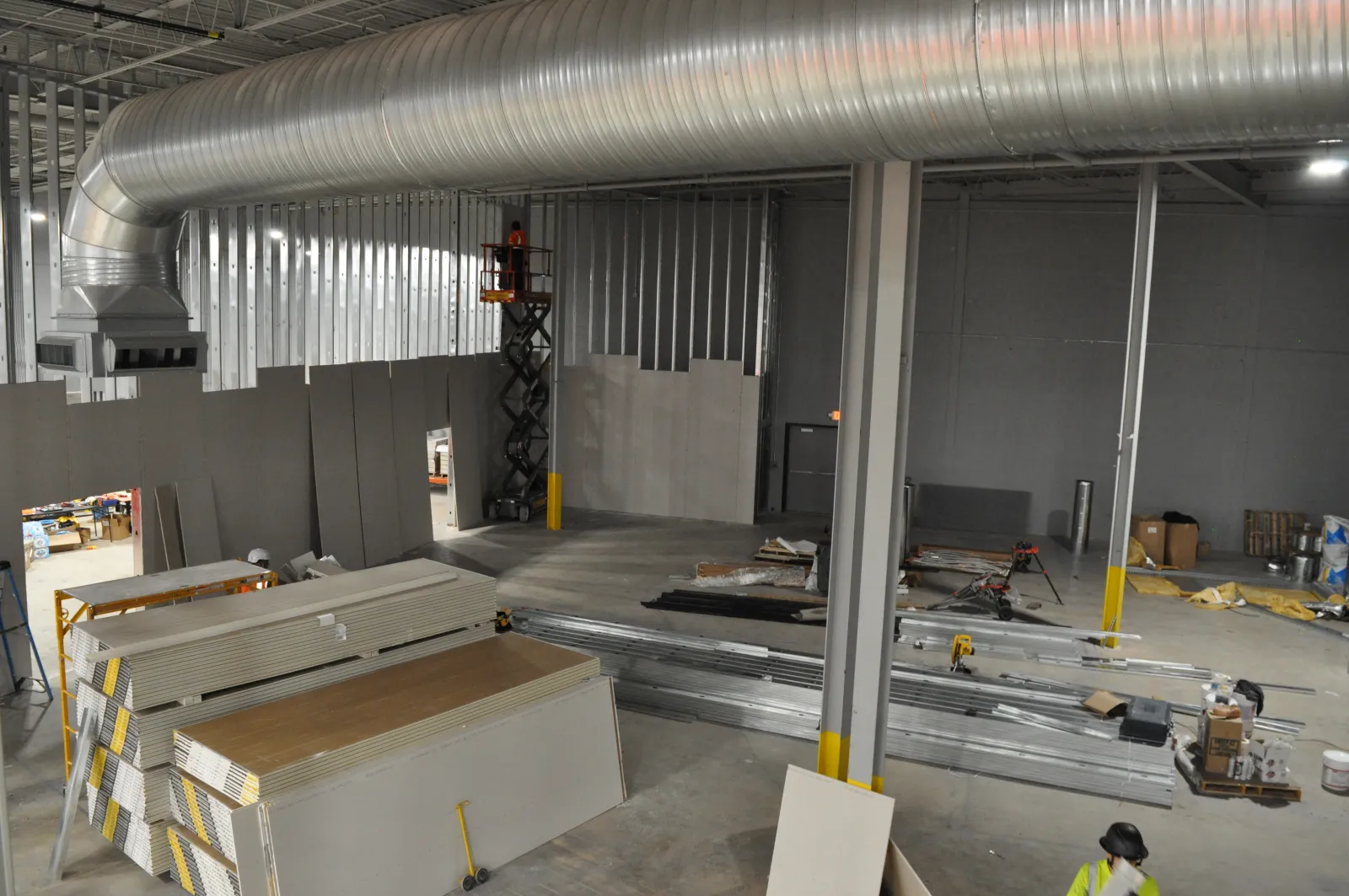 Interior view of a warehouse under construction with drywall stacks, steel beams, and a worker in safety gear.