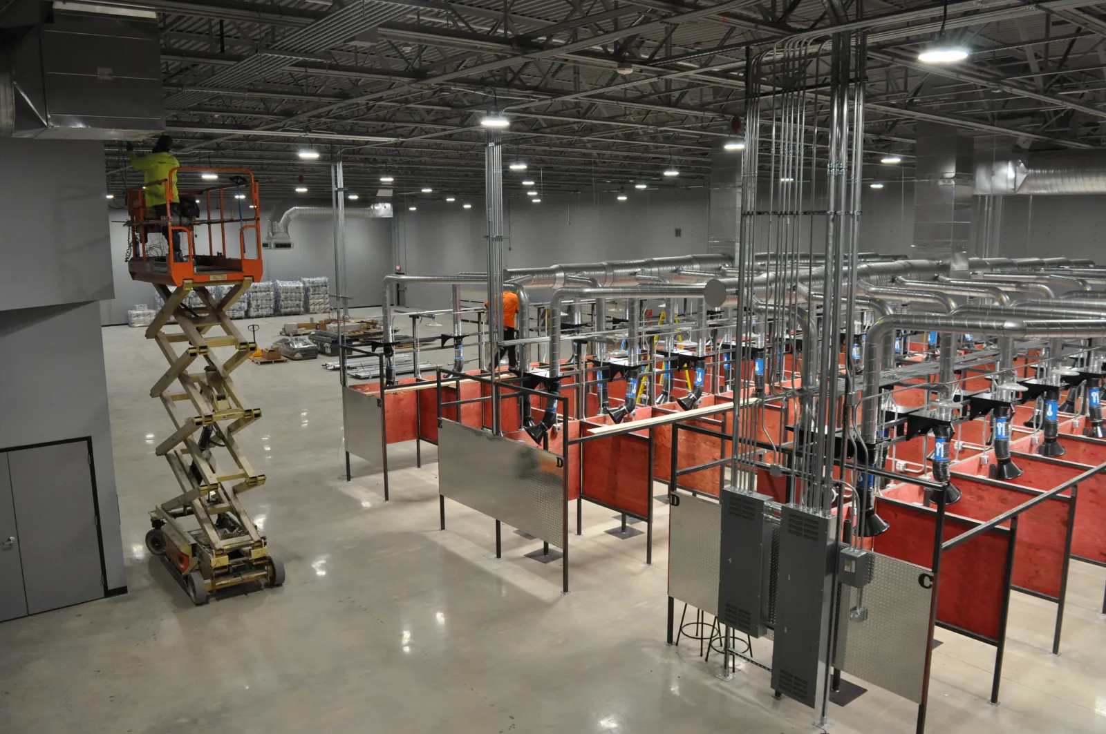 Worker on scissor lift in a large industrial welding training facility with ventilation and red partitions.