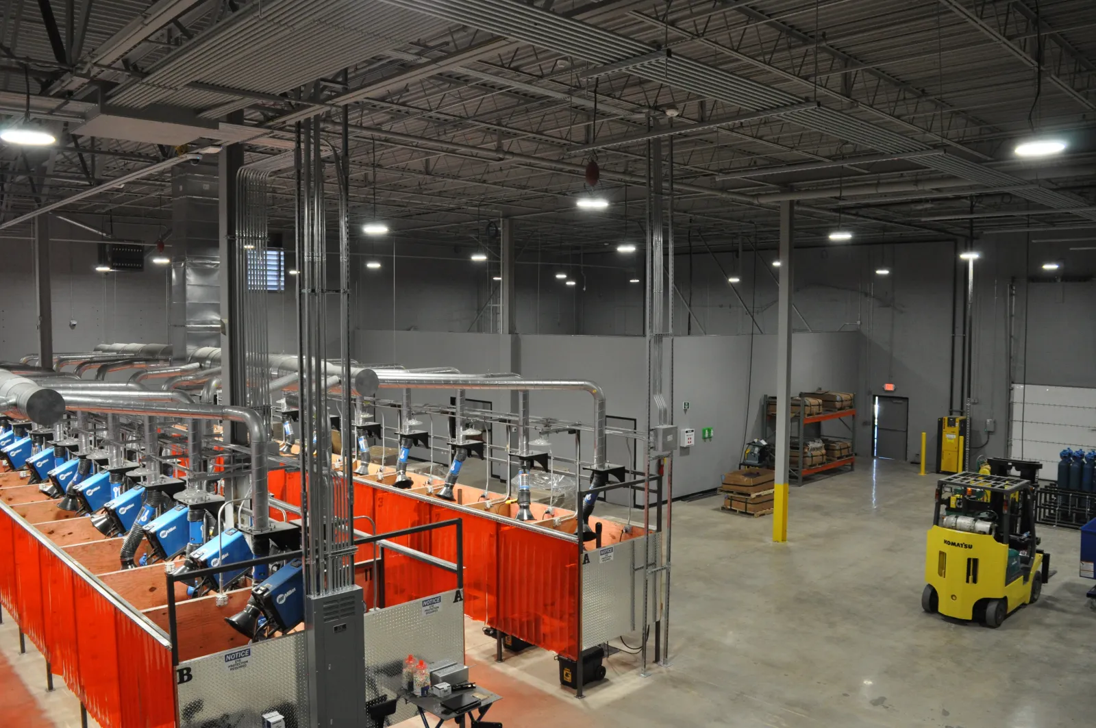 Spacious industrial warehouse with rows of red welding stations and a yellow forklift under high ceiling lights.