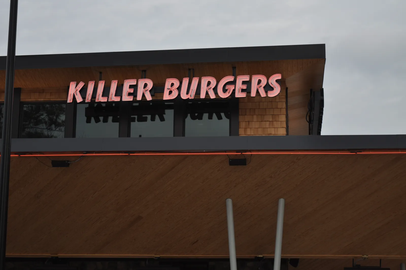 Killer Burgers neon sign on a modern wooden and glass restaurant facade during overcast weather.