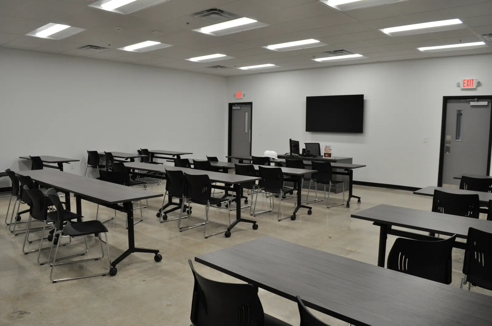 Modern empty classroom with black chairs, gray tables, concrete floor, and a large monitor on white wall.