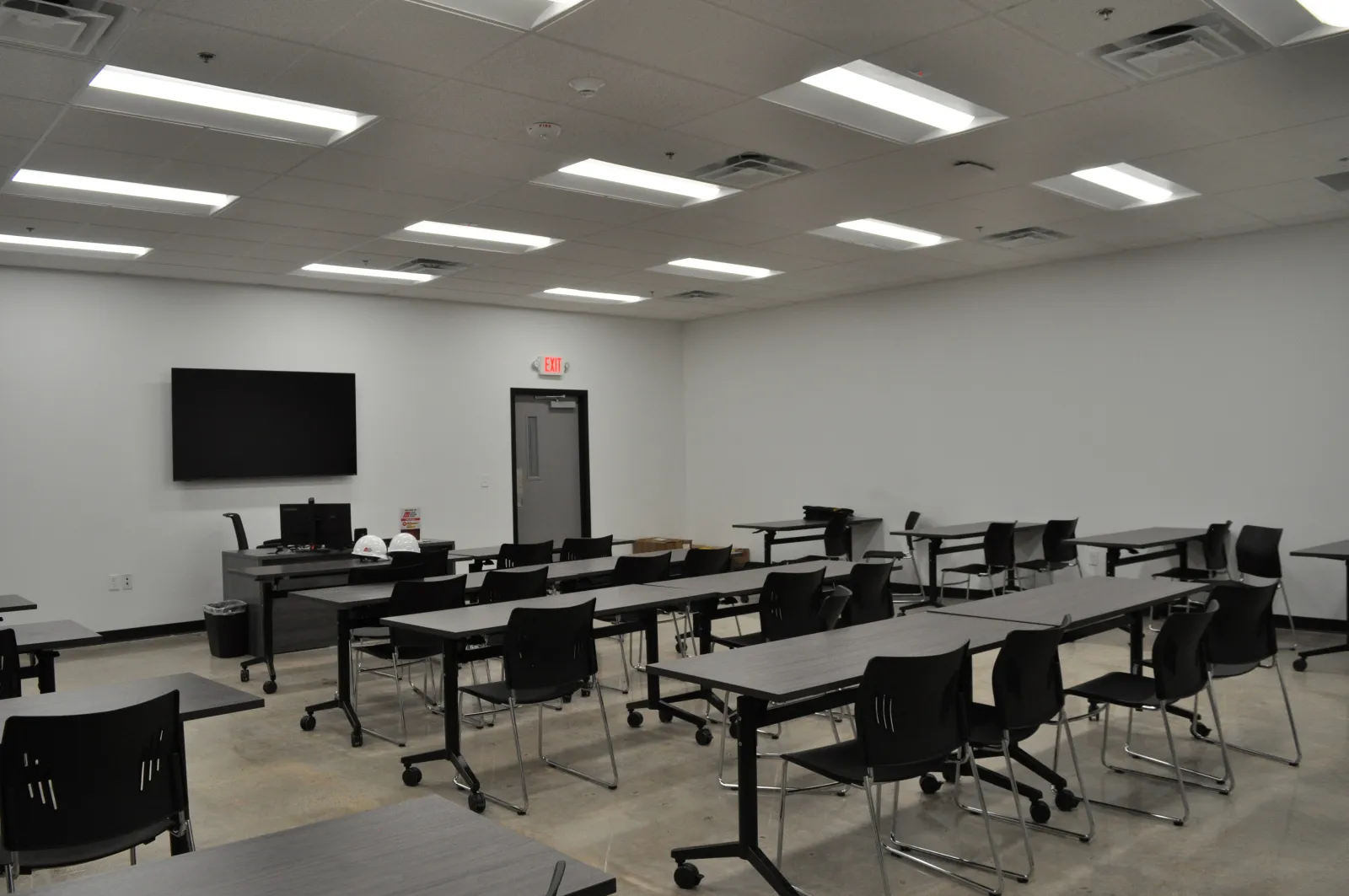 Empty modern classroom with black chairs, gray tables, wall-mounted screen, and fluorescent ceiling lights.