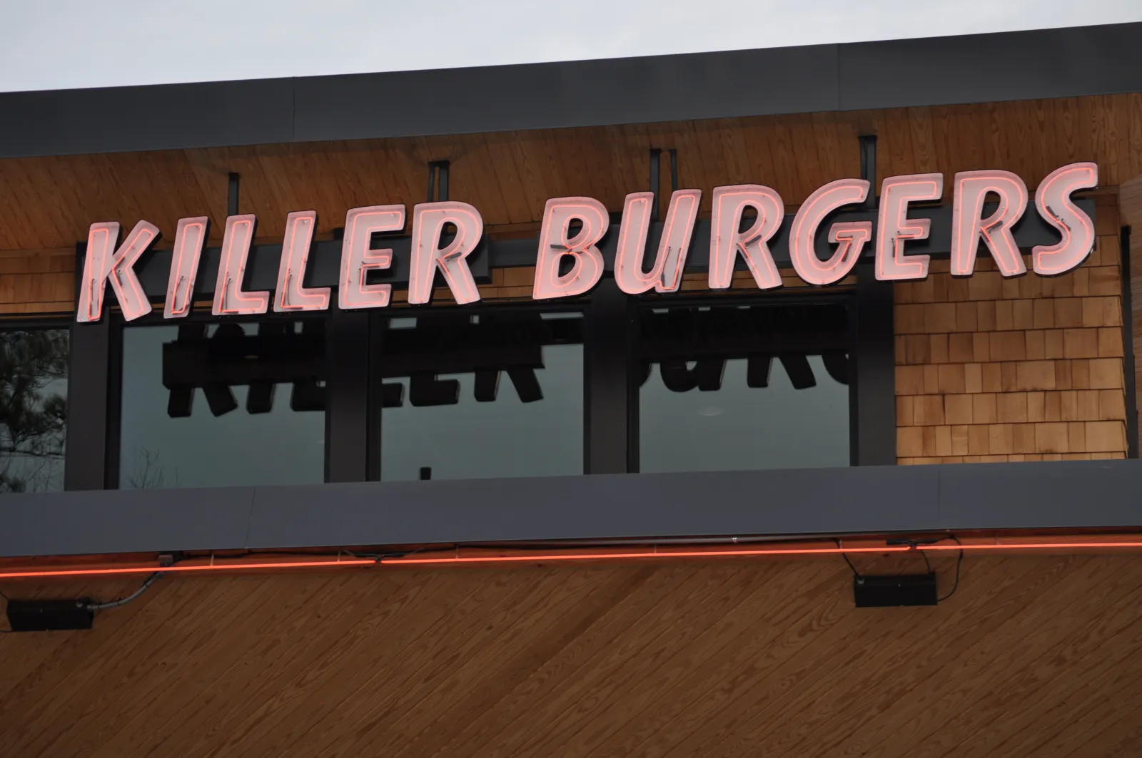 Killer Burgers neon sign mounted on wooden shingle wall above windows at burger restaurant.