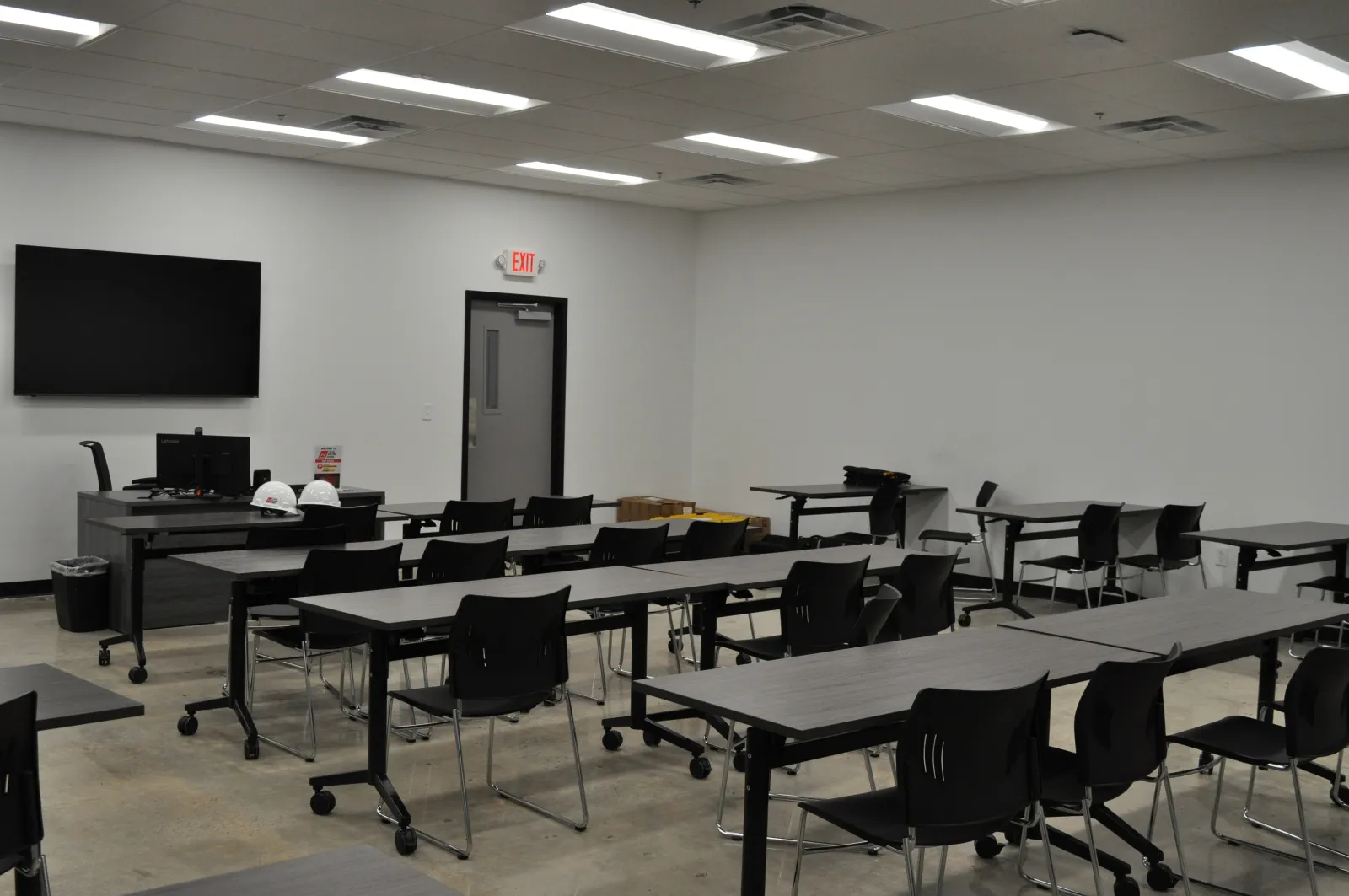 Modern empty classroom with black chairs, rectangular tables, a large screen, and white walls under fluorescent lights.