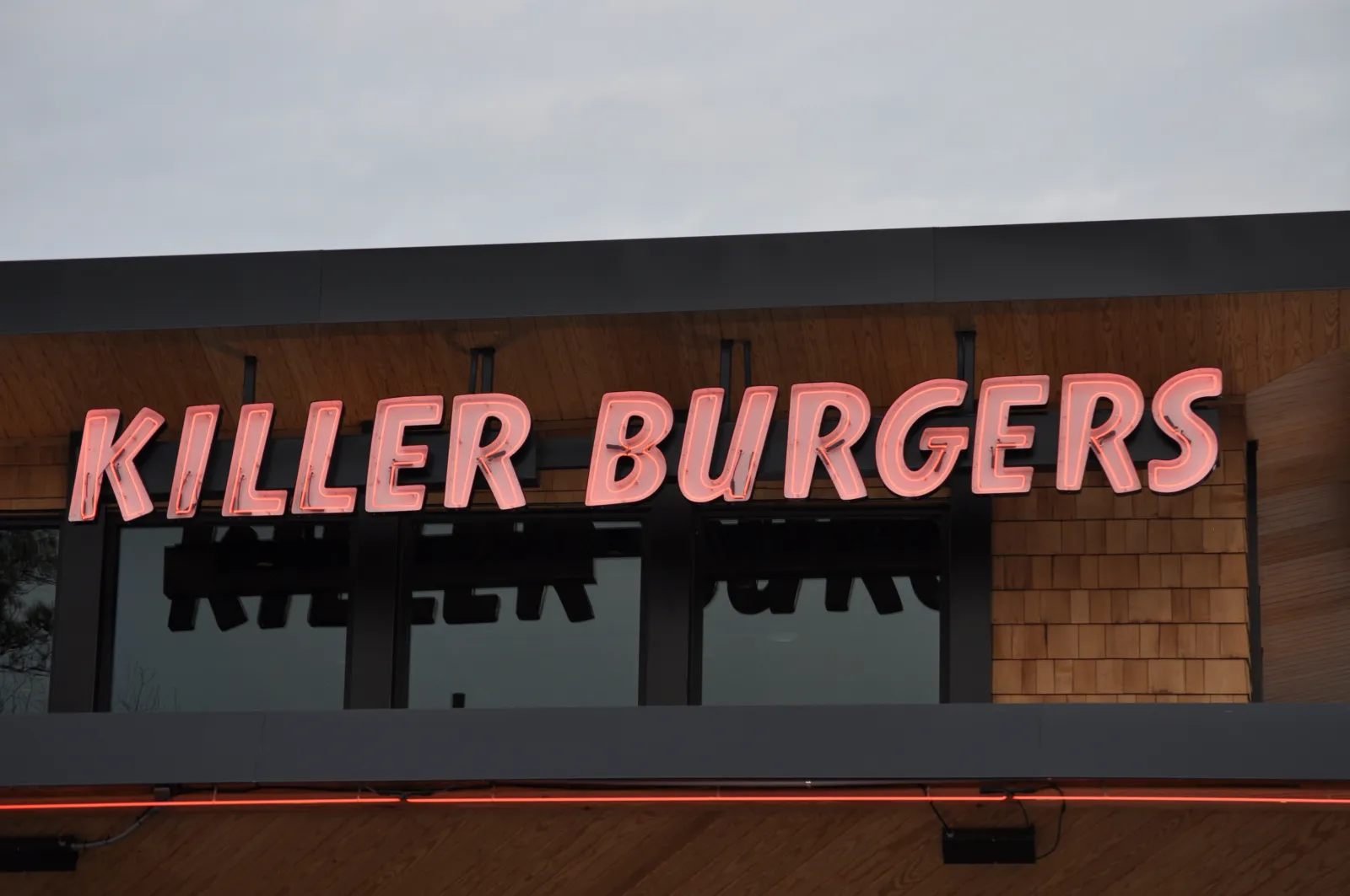 Neon pink sign reading Killer Burgers mounted on modern wooden and black storefront under cloudy sky.