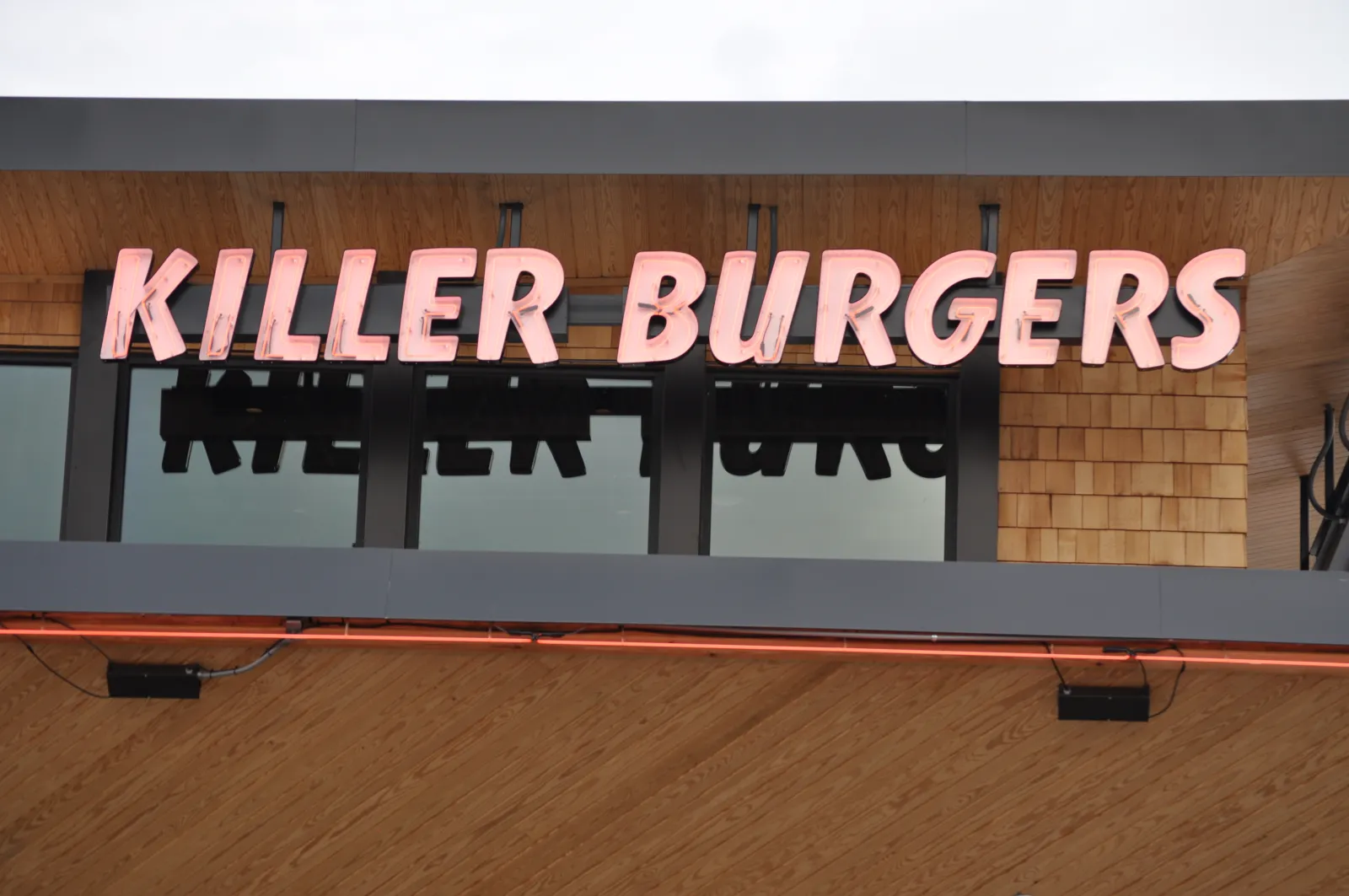 Signage of Killer Burgers restaurant with illuminated letters on wooden and glass facade.