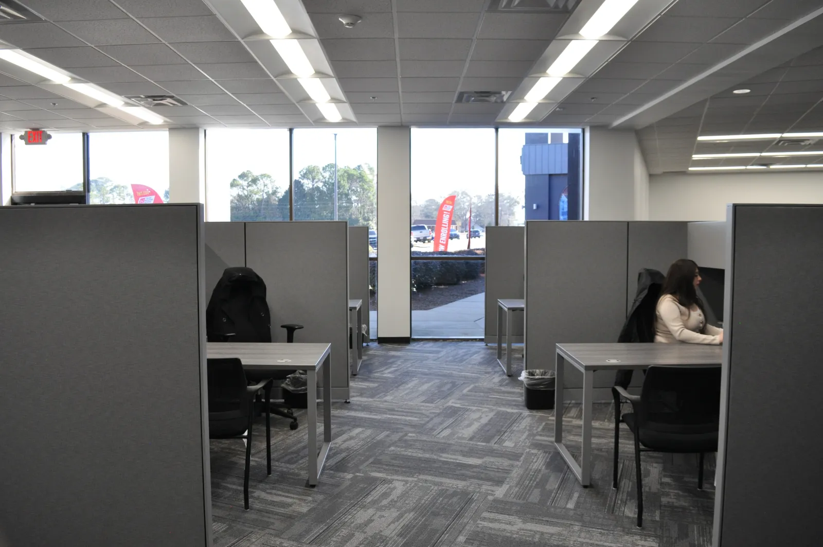 Office cubicles with gray partitions and desks, a woman working on a computer by the window, bright natural light.