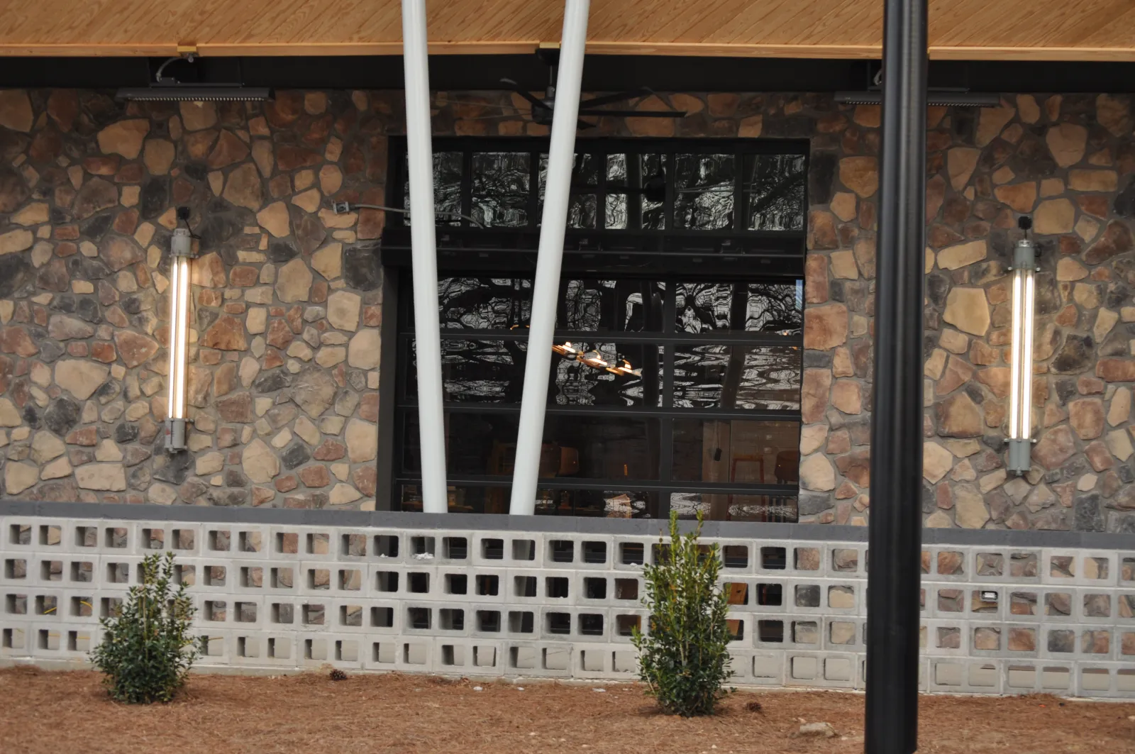 Stone wall exterior with large window, modern vertical lights, and small bushes in front under wooden overhang.