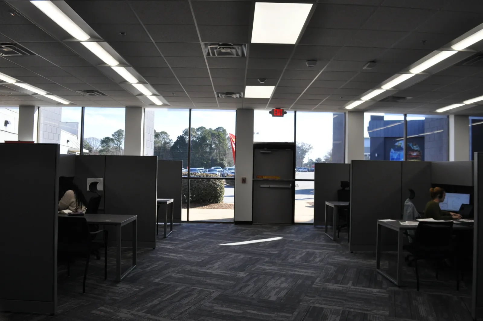 Modern office space with cubicles, desks, computers, and large windows showing outdoor parking lot.