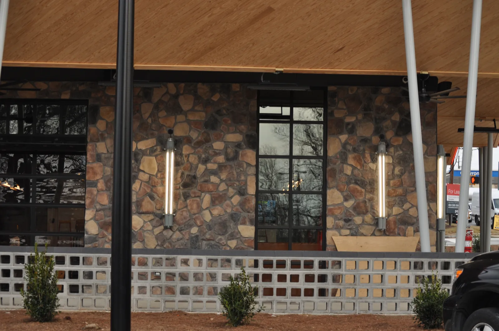 Stone exterior wall with tall windows and modern vertical lights under a wooden roof with small shrubs.
