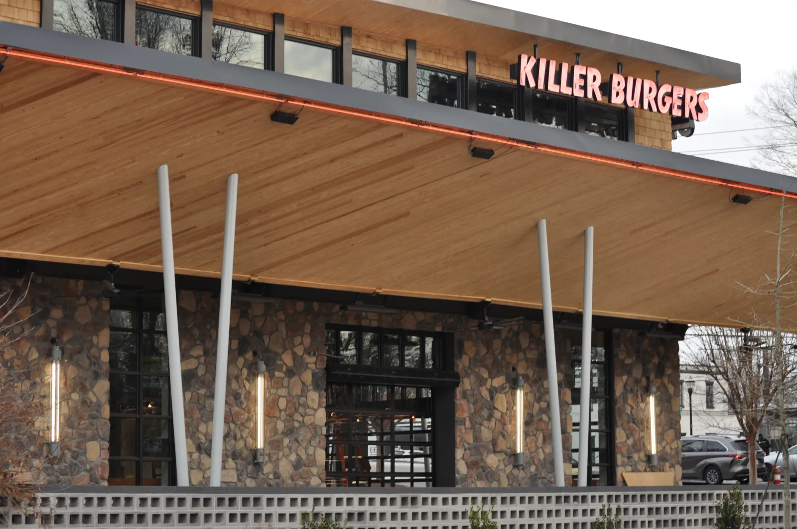 Exterior view of Killer Burgers restaurant with stone facade, wooden canopy, and illuminated vertical lights.