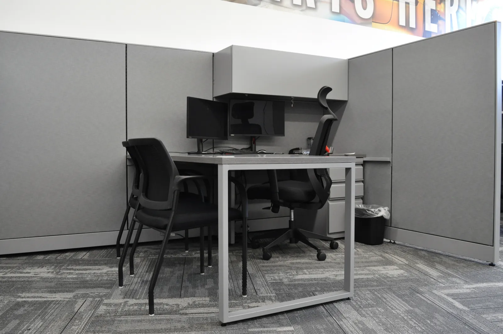 Modern office cubicle with gray walls, two black chairs, dual monitors, and a desk with gray metal frame.