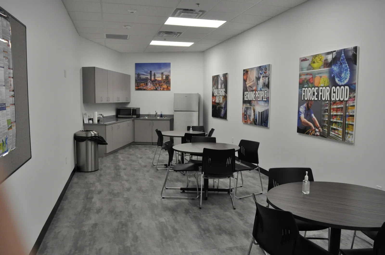 Corporate break room with round tables, black chairs, kitchenette, refrigerator, and motivational posters on walls.
