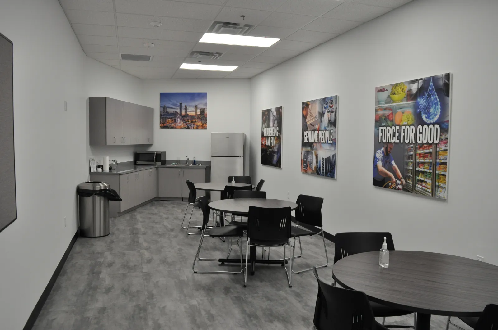Modern office break room with kitchen area, round tables, black chairs, motivational posters, and a gray floor.