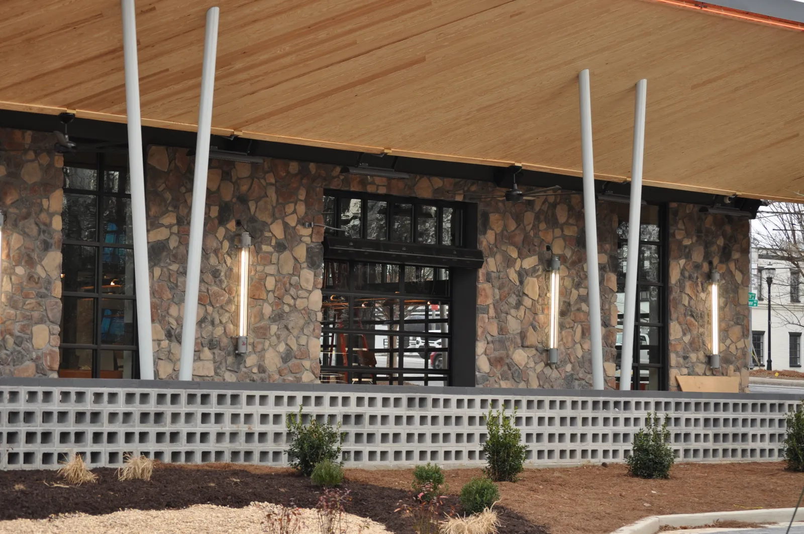 Modern building exterior with stone wall, large windows, vertical lights, and wooden roof overhang.