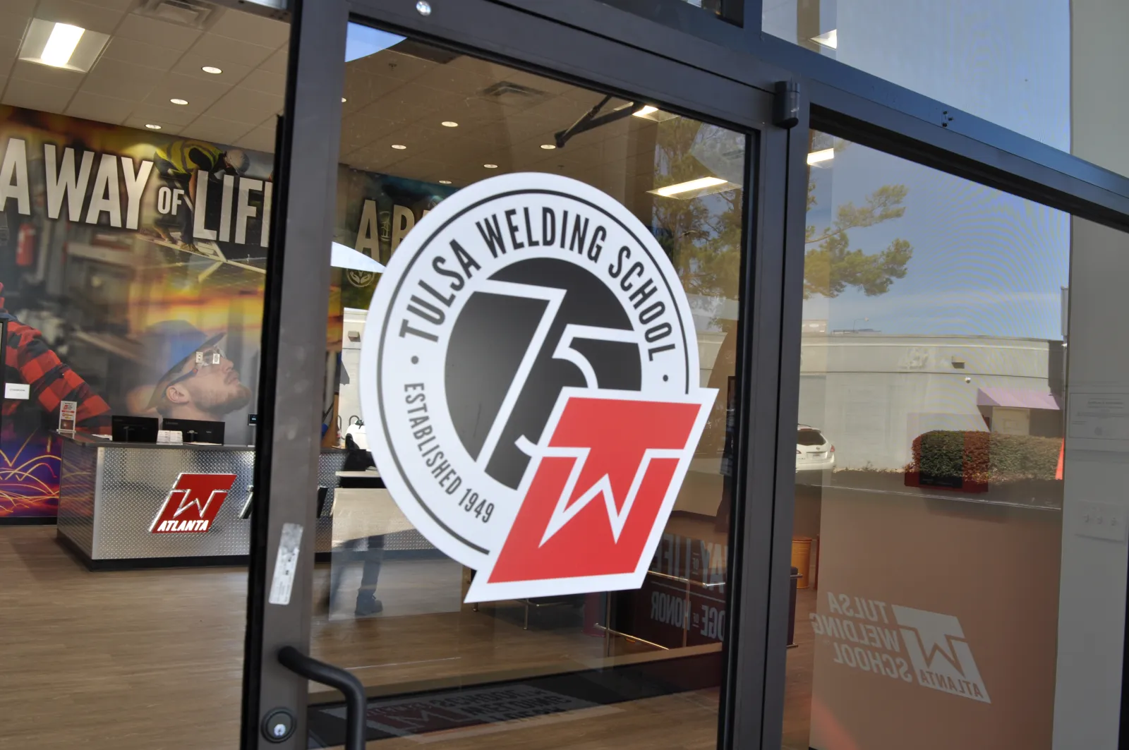 Entrance glass door with Tulsa Welding School logo and interior showing workspace and wall art