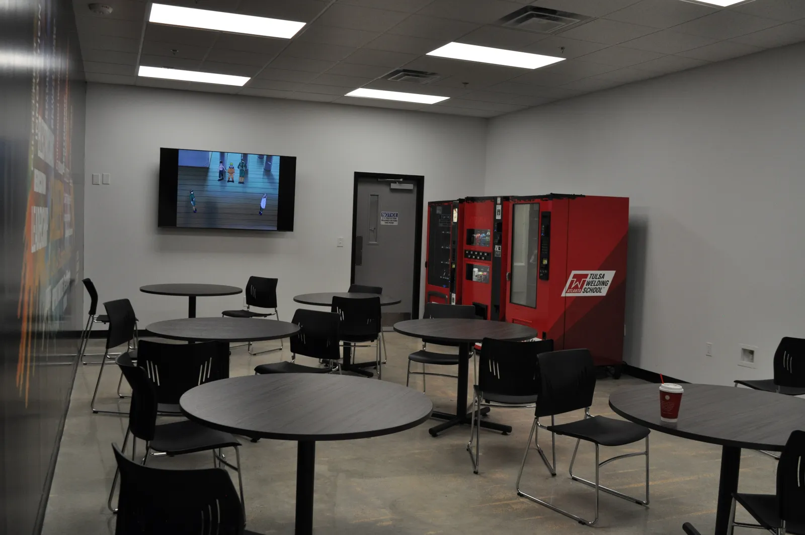 Empty break room with round tables, black chairs, red vending machines, and a TV mounted on a white wall.