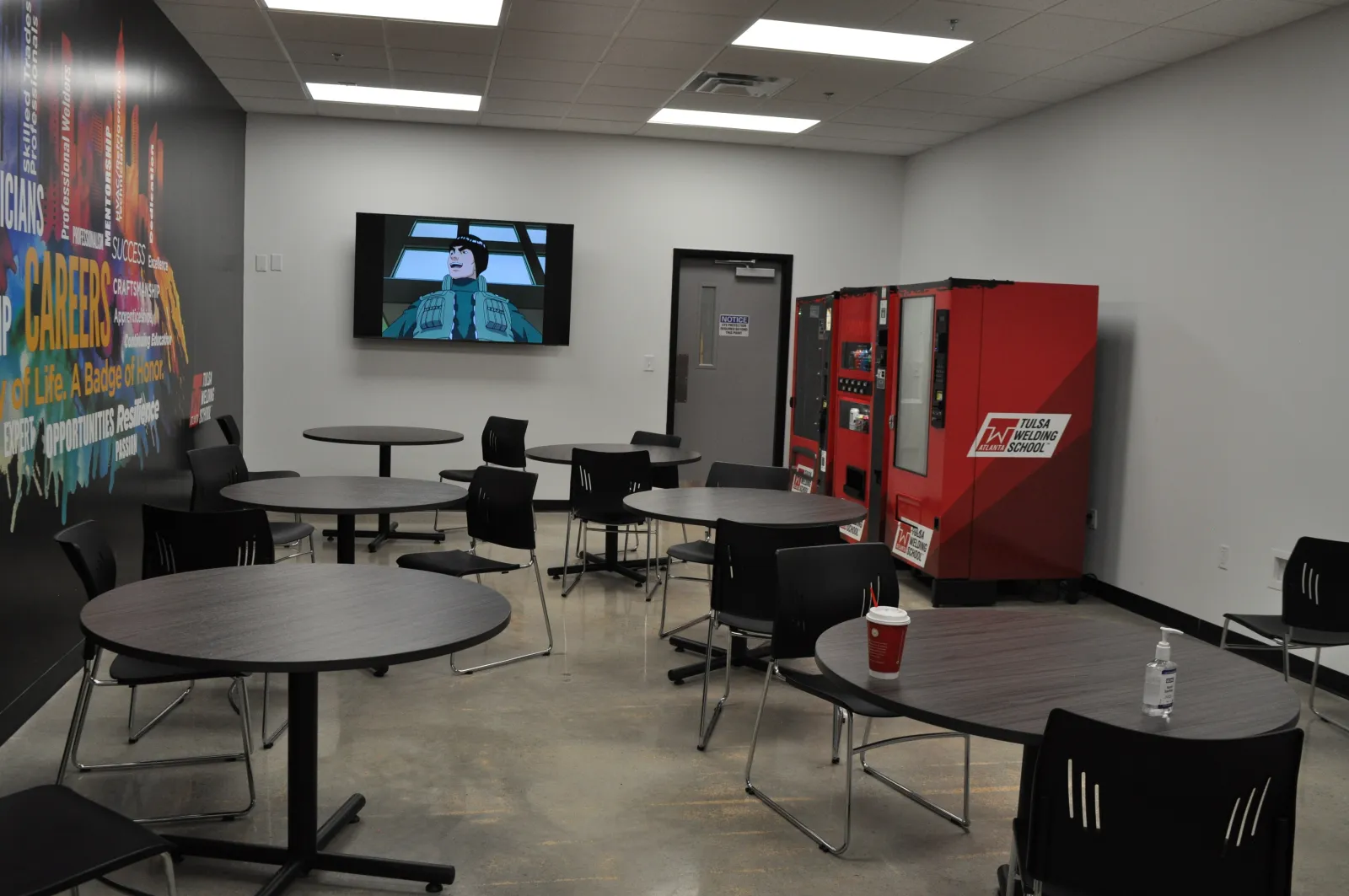 Modern break room with round tables, black chairs, vending machines, TV screen, and motivational wall graphics.