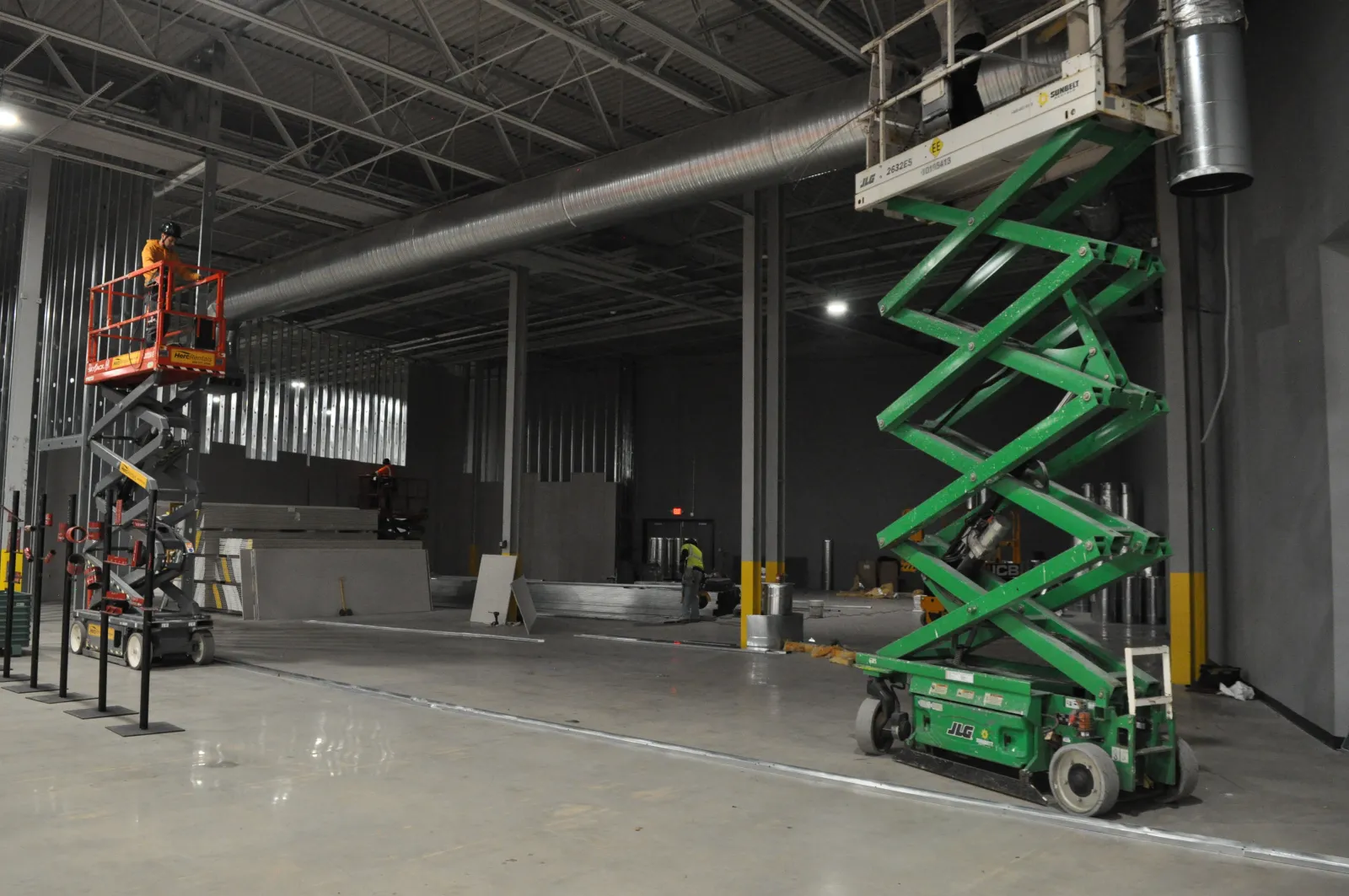 Workers using scissor lifts inside a large industrial warehouse under construction with metal framing and concrete floors