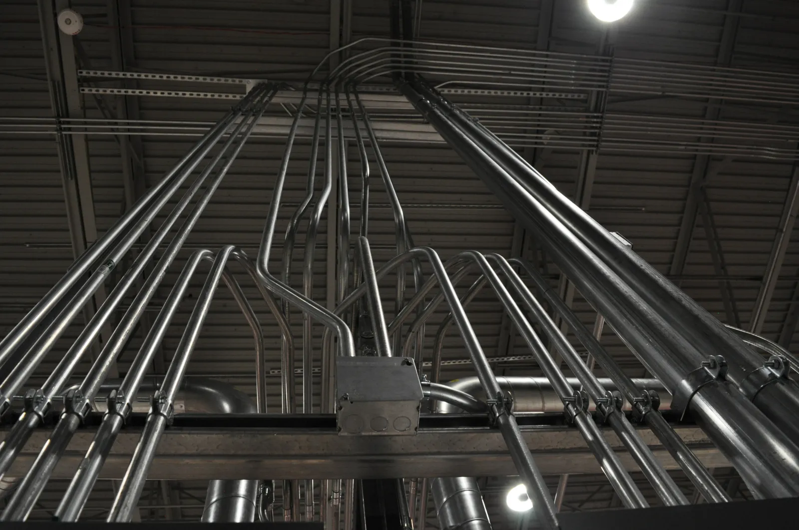 Multiple metal electrical conduits mounted on ceiling beams in an industrial setting.