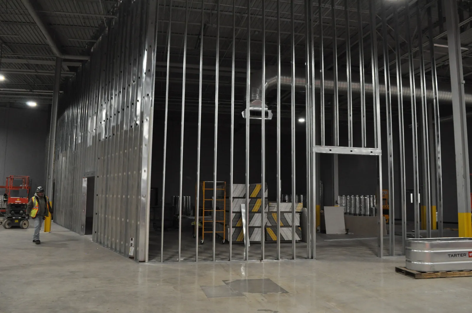 Construction site interior with metal stud framing for walls, stacks of drywall, and a worker in safety vest.