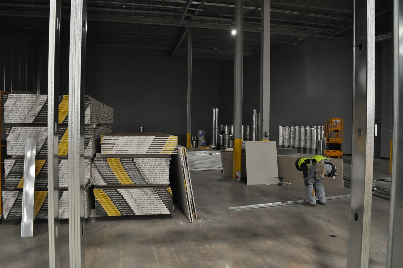 Worker installing drywall panels inside a large, unfinished warehouse with metal framing and construction materials.