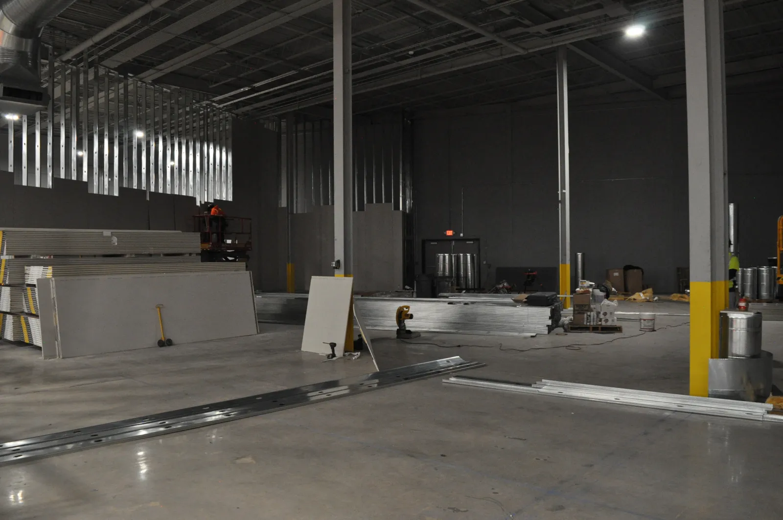 Large warehouse interior under construction with drywall sheets, metal beams, and construction tools scattered.
