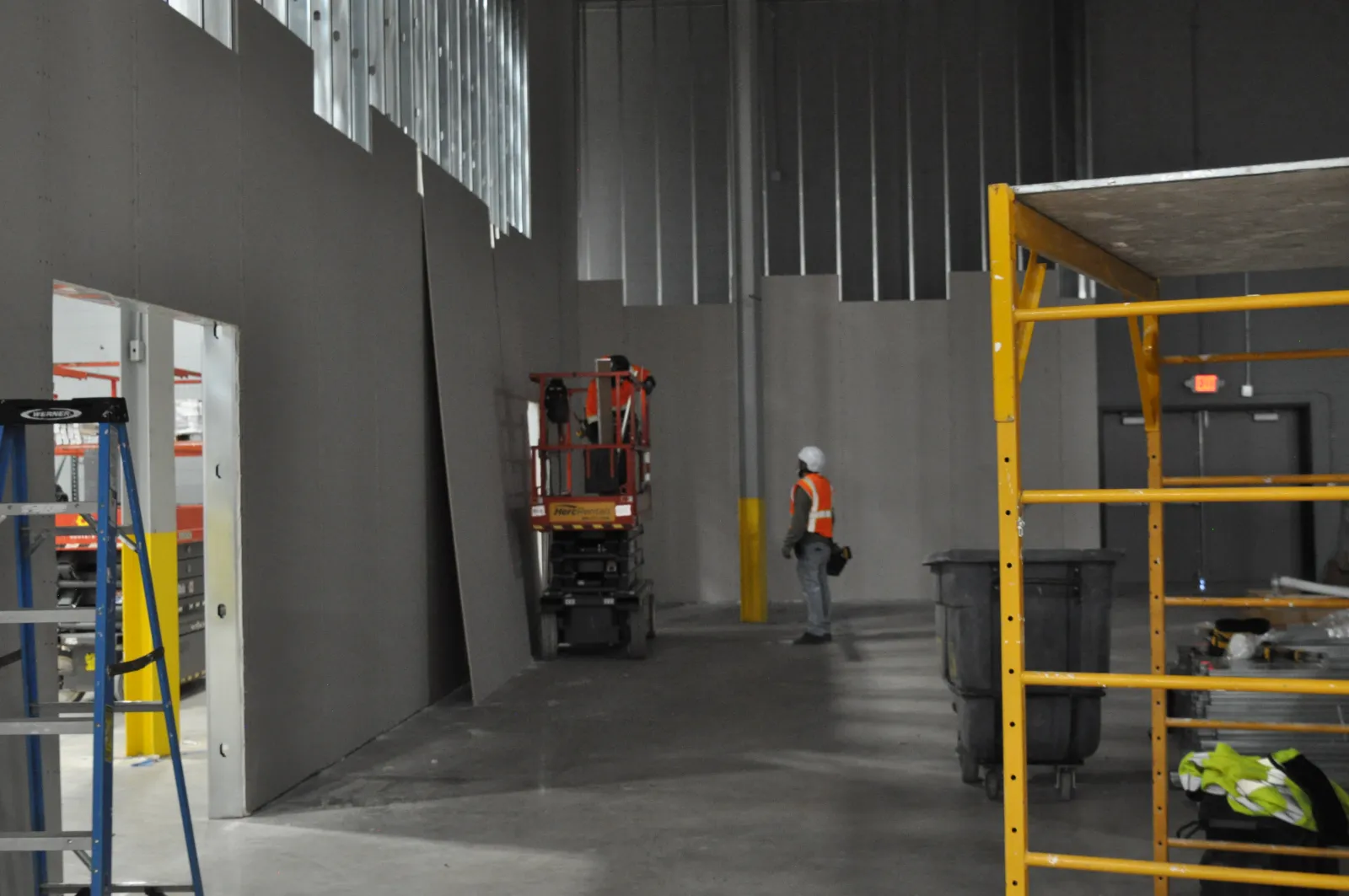 Construction worker in safety vest and helmet inside large unfinished industrial building with scaffolding and equipment.