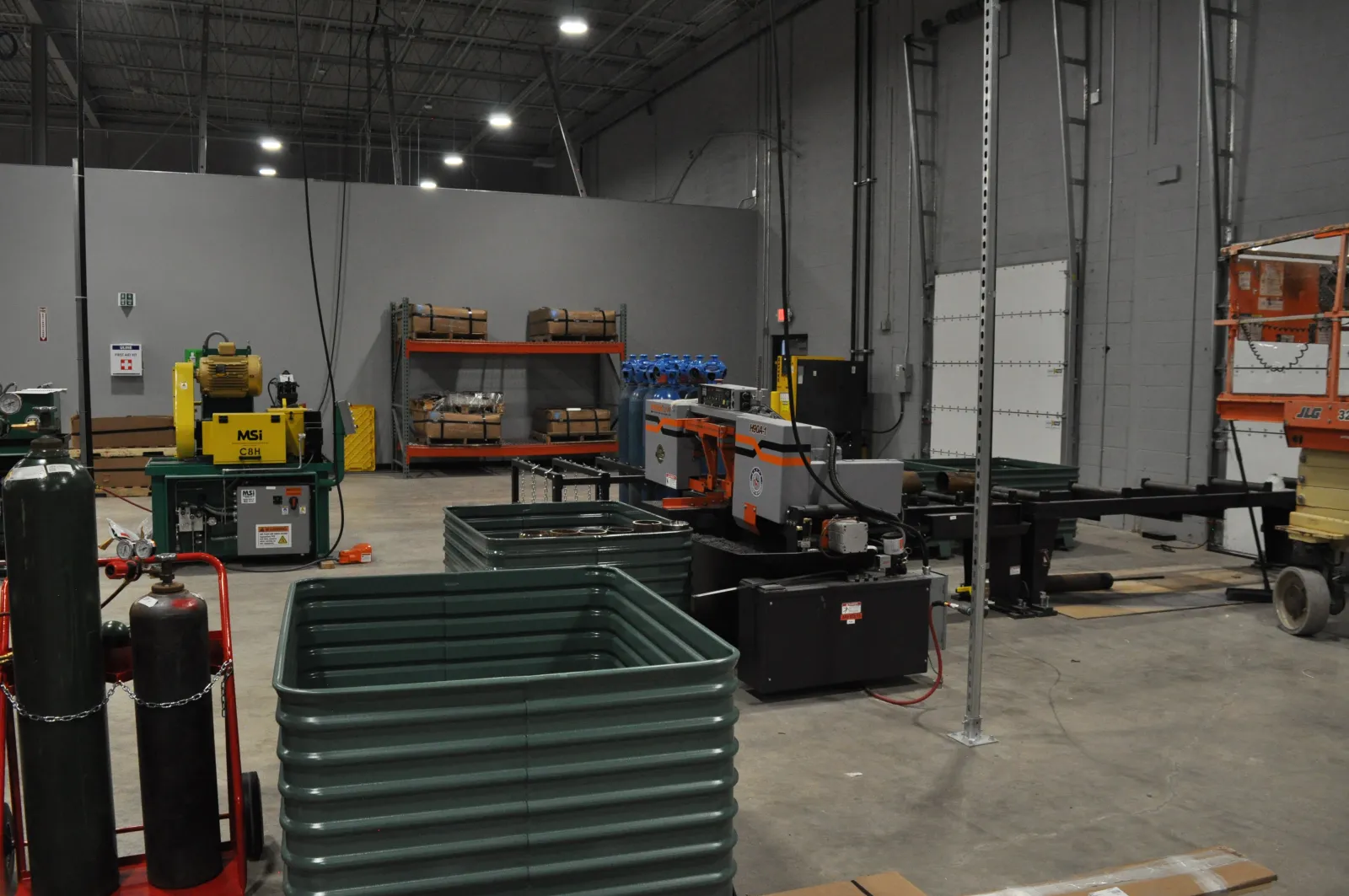 Industrial warehouse interior with metal machinery, stacked materials, and equipment on concrete floor under high ceiling.