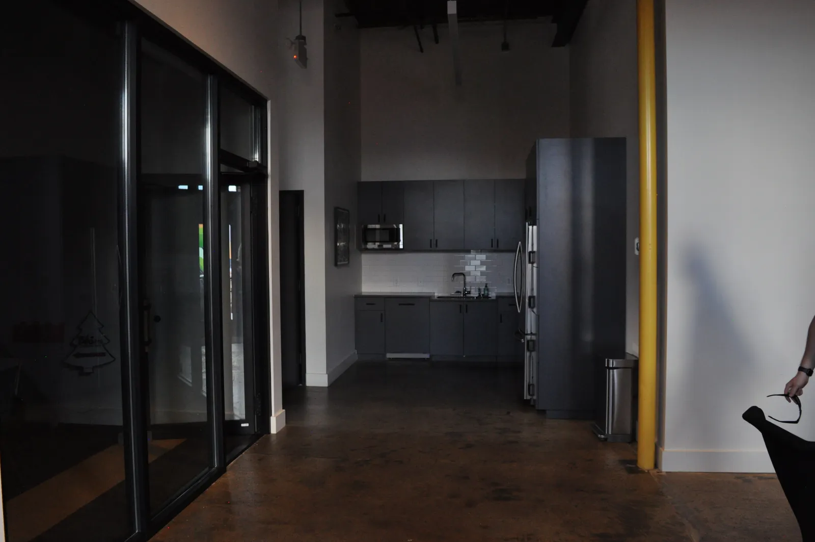 Modern office kitchen area with dark cabinets, stainless steel appliances, and polished concrete floor under dim lighting