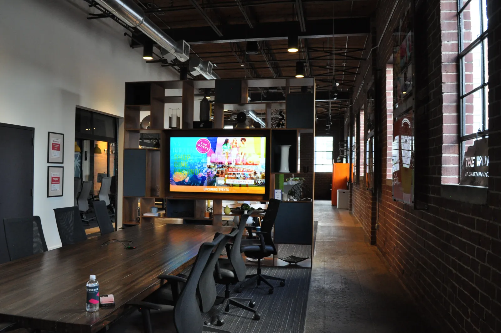 Modern conference room with long wooden table, office chairs, large screen, and brick walls in industrial space.