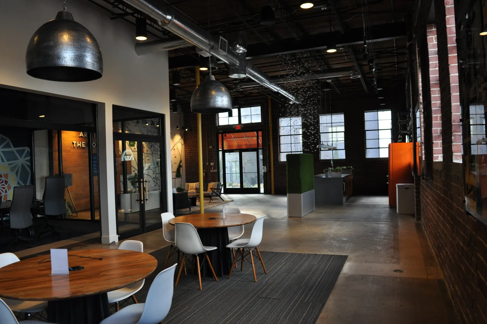 Modern industrial office space with round wooden tables, white chairs, exposed brick walls, and large windows.