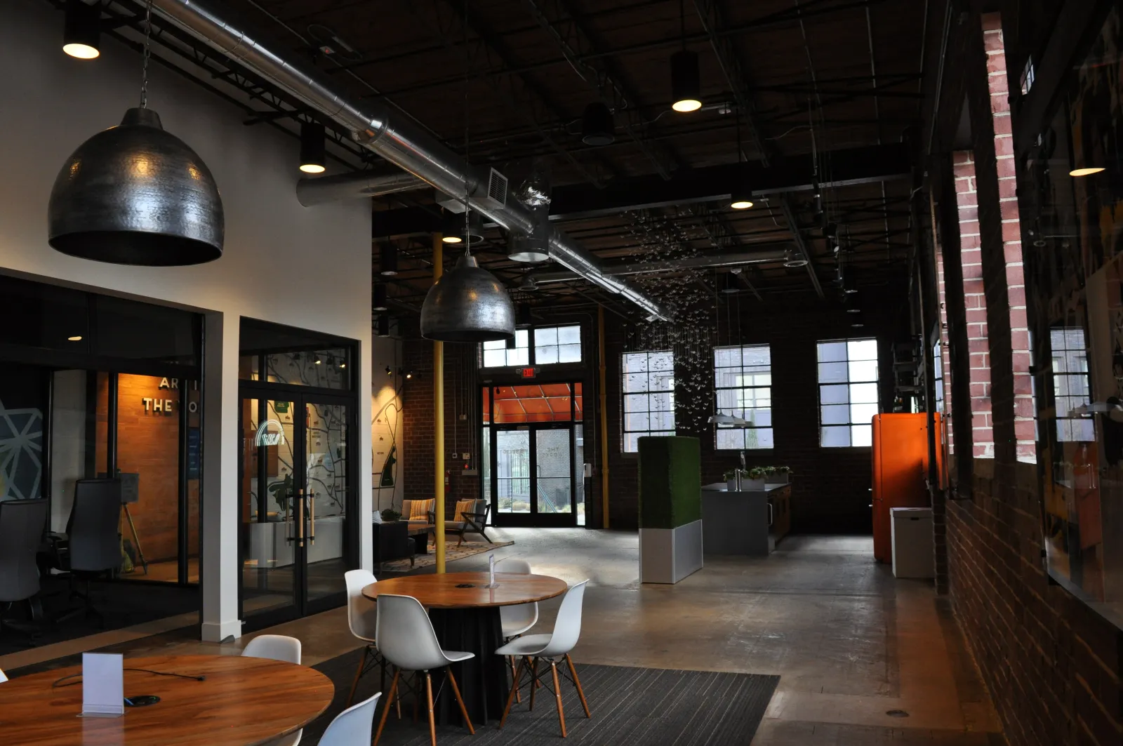 Modern office space with round wooden tables, white chairs, industrial lighting, and large windows.