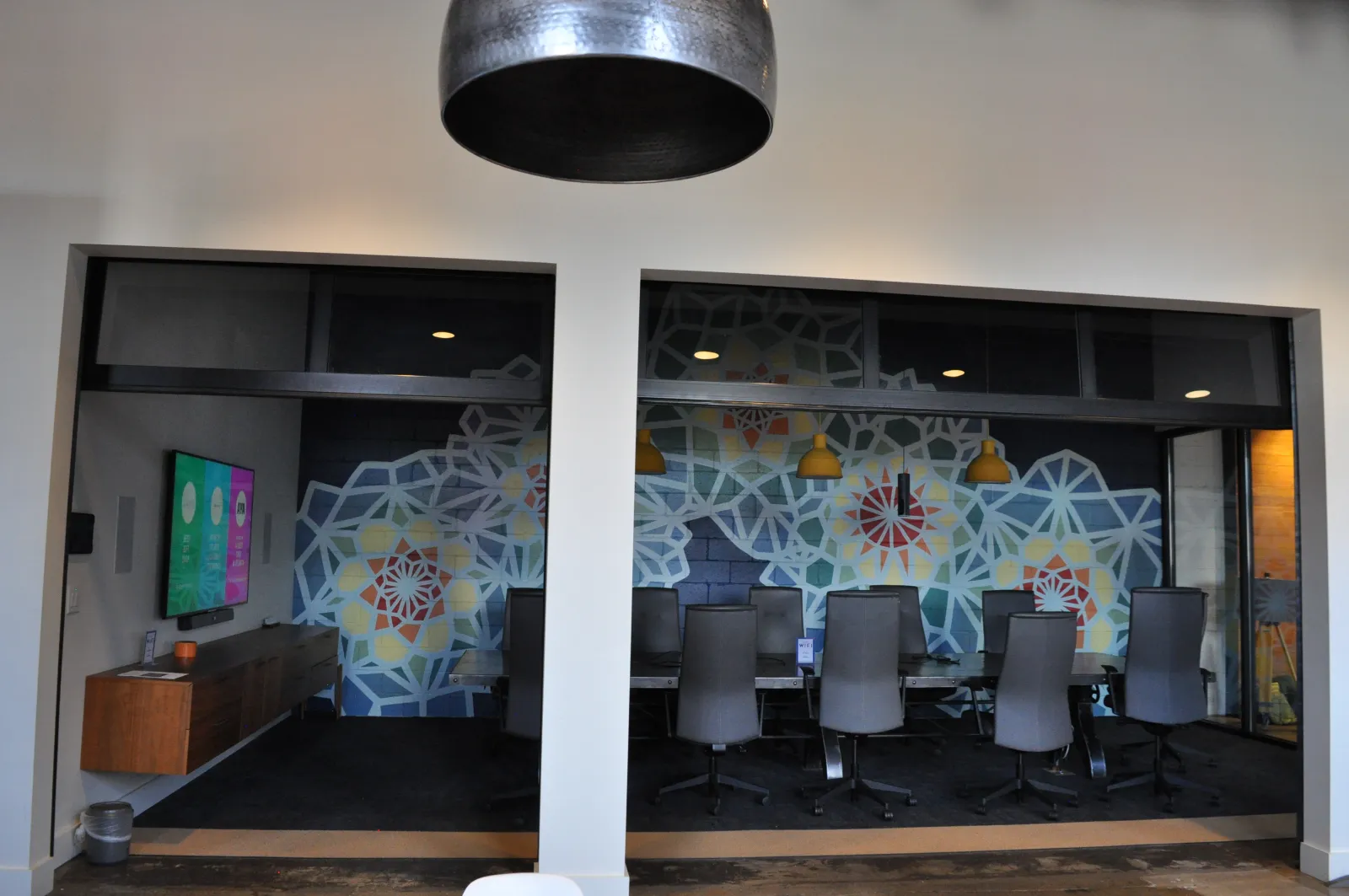 Modern conference room with patterned wall art, black chairs, pendant lights, and a mounted flat screen TV.
