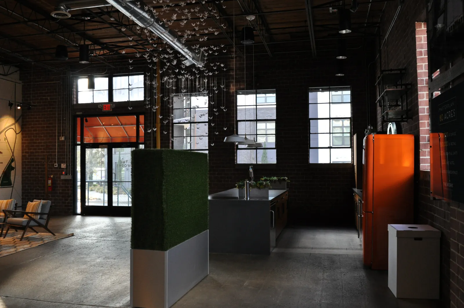 Industrial-style office interior with brick walls, large windows, orange refrigerator, and modern furniture.
