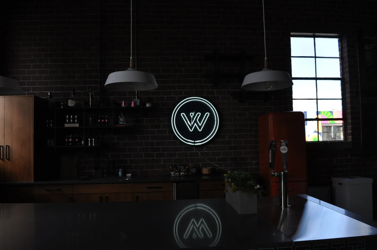 Dark industrial kitchen with brick walls, hanging lights, neon W logo, and orange refrigerator by a window.