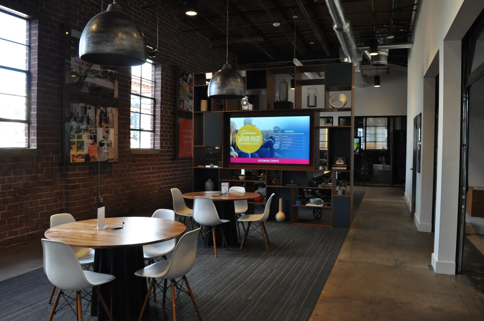 Modern coworking space with round tables, white chairs, pendant lights, and a digital screen on a bookshelf.