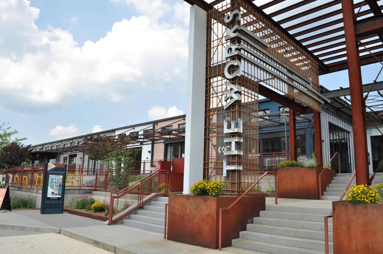 Exterior of a modern building with rust-colored metal structure and stairs under a cloudy sky.