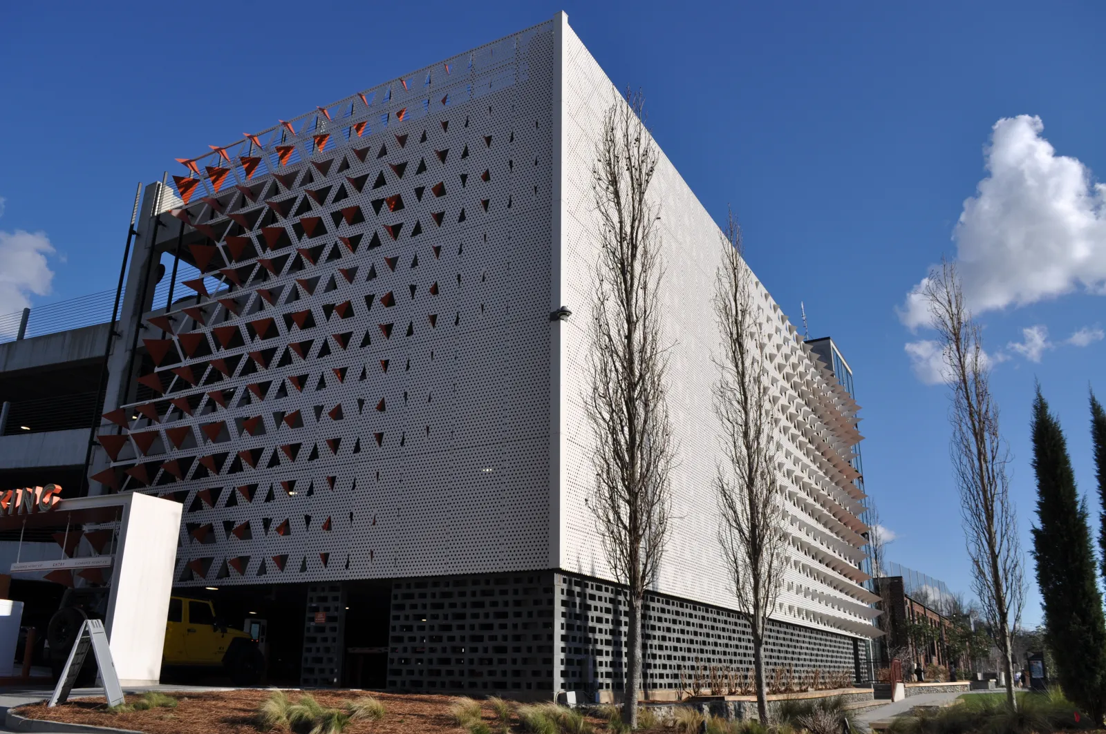 Modern multi-story parking garage with perforated metal facade and tall trees under clear blue sky