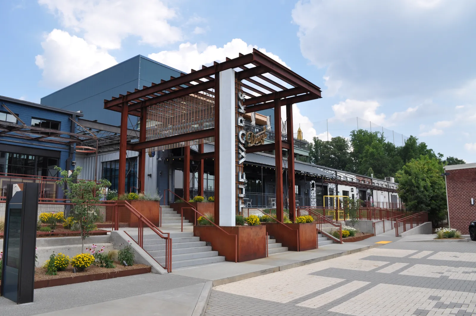 Modern building entrance with rust-colored steel pergola, concrete stairs, and landscaped planters under a partly cloudy sky