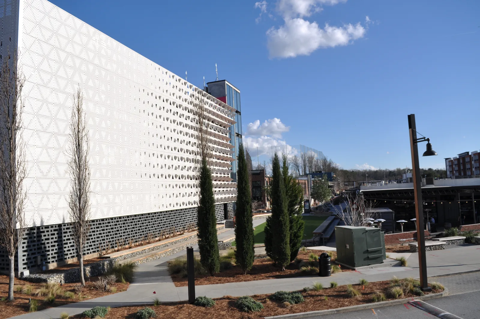 Modern building with perforated white facade, tall trees, pathways, and clear blue sky in an urban landscape.