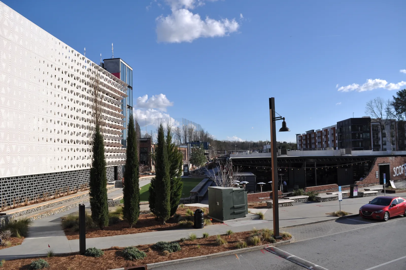 Modern urban street scene with trees, contemporary buildings, clear sky, and parked red car on a sunny day.