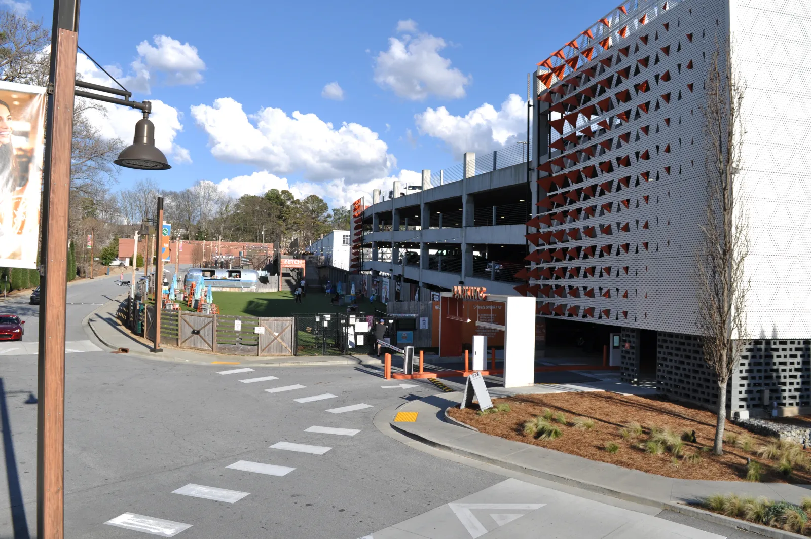 Modern multi-story parking garage beside a street with a fenced playground, trees, and a partly cloudy sky.