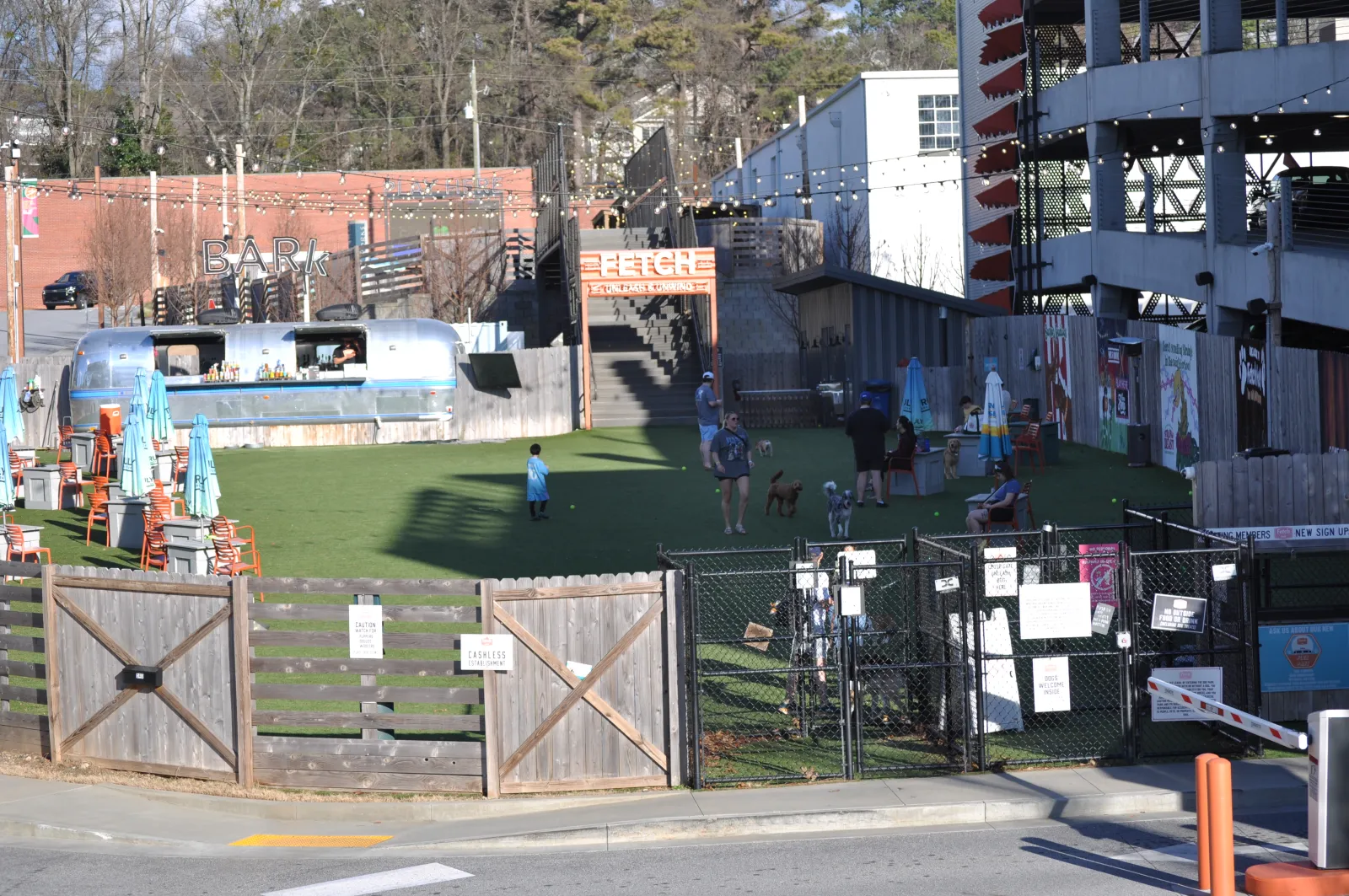 Outdoor dog park with fenced area, grass field, people, dogs, and a mobile bar in an urban setting on a sunny day