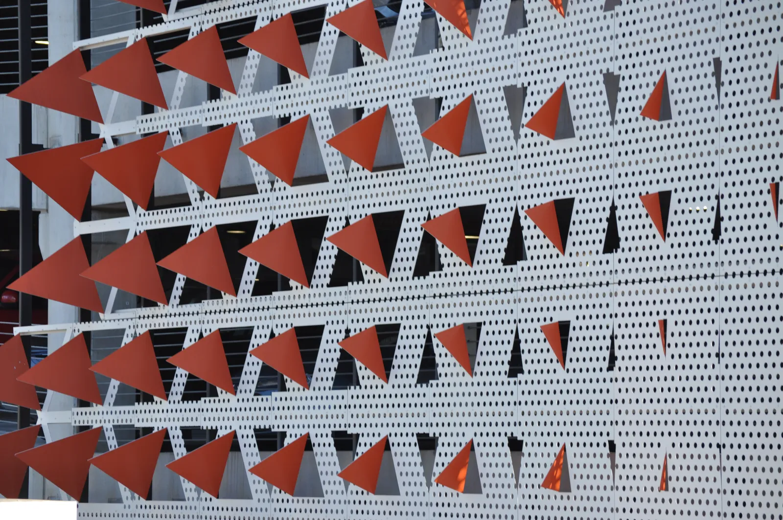 Facade with rows of red triangular metal elements mounted on perforated white panels of a building exterior.