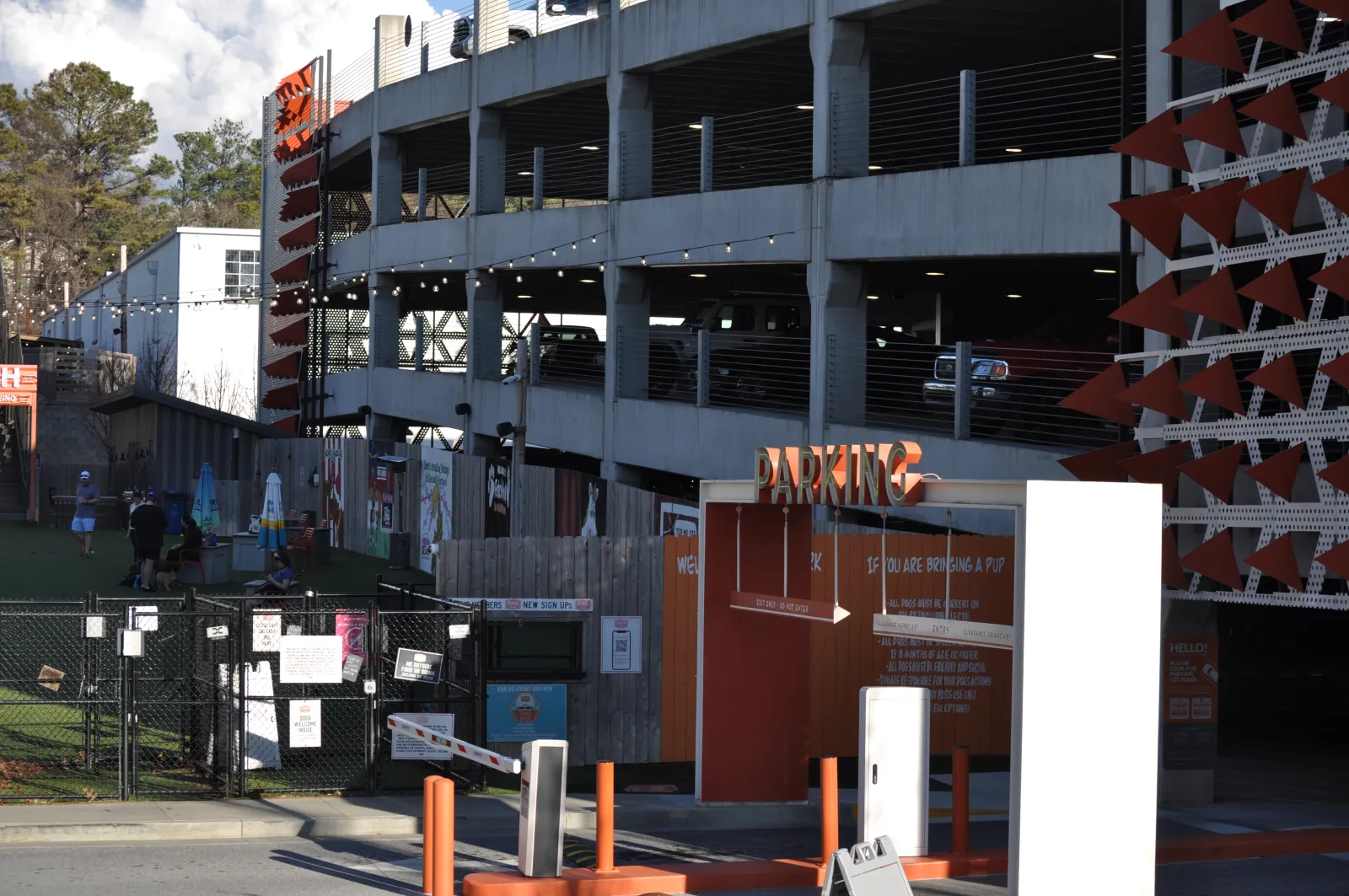 Outdoor parking garage entrance with orange signage and nearby green fenced area under partly cloudy sky.