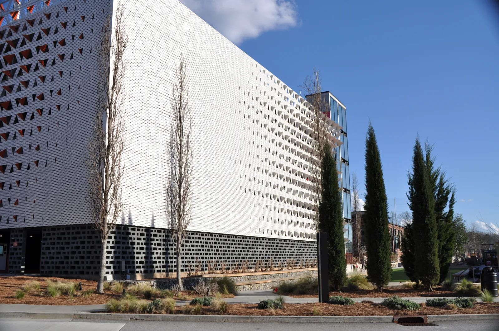 Modern building with perforated white facade and black base surrounded by tall trees on a sunny day.