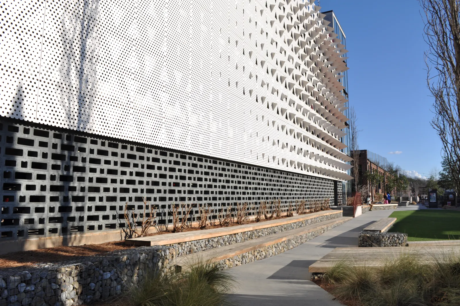 Modern building facade with perforated white panels, geometric patterns, and landscaped outdoor seating area under clear sky.