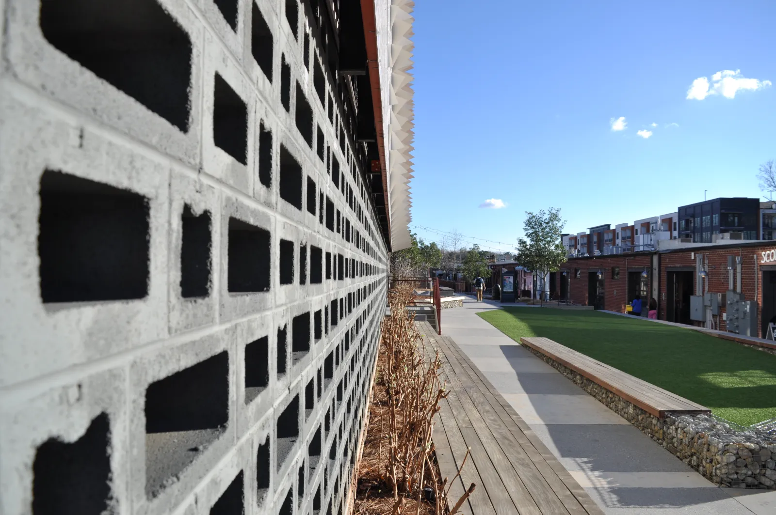 Modern outdoor space with patterned concrete wall, wooden benches, green lawn, and urban buildings under blue sky.
